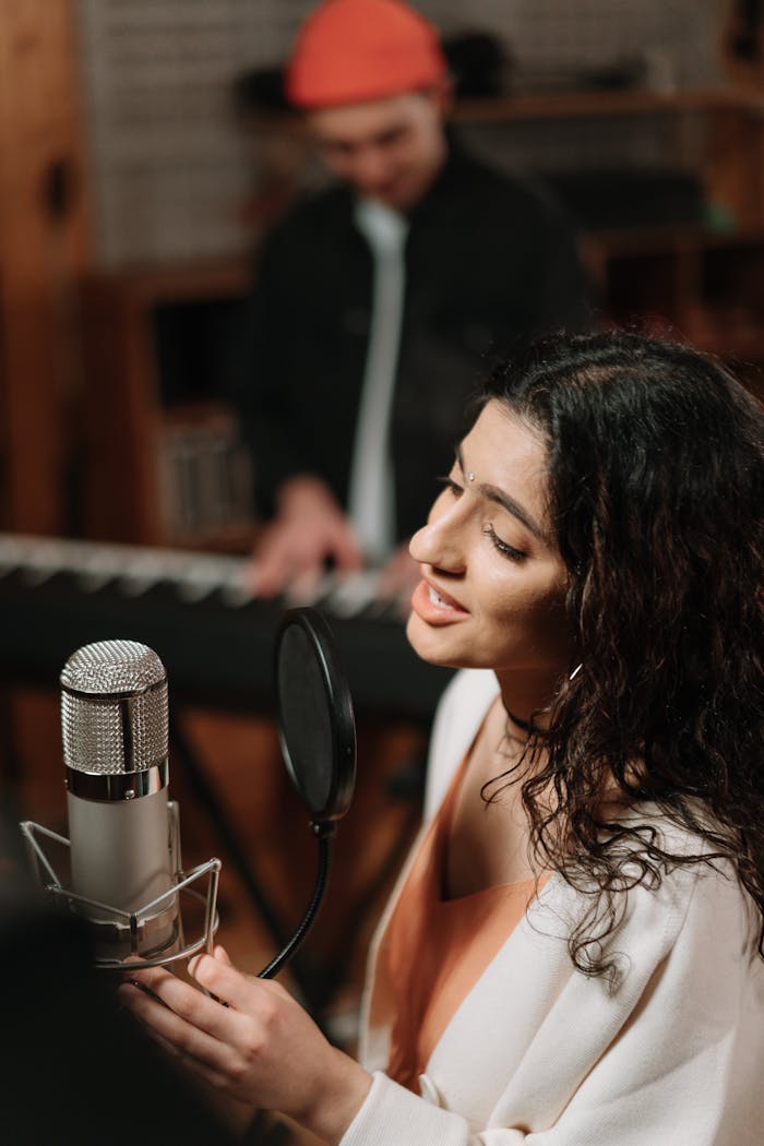 About Woman singing into microphone with musician at keyboard in a recording studio.